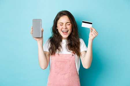 兴奋的漂亮女人欢呼雀跃，展示空的智能手机屏幕和塑料信用卡，举起双手，快乐地微笑，站在蓝色背景上