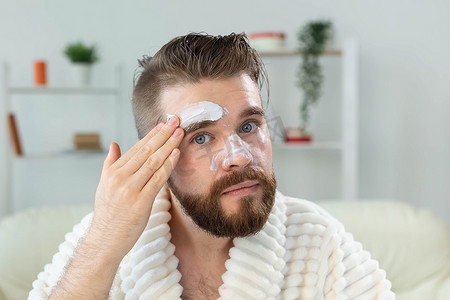 男士护肤摄影照片_留胡子的男人在镜子前涂面霜 护肤和水疗为男士概念。