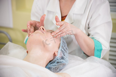 在诊所为一位闭着眼睛的年轻女性做面部按摩的特写镜头，医生的手抚摸着病人的脸颊