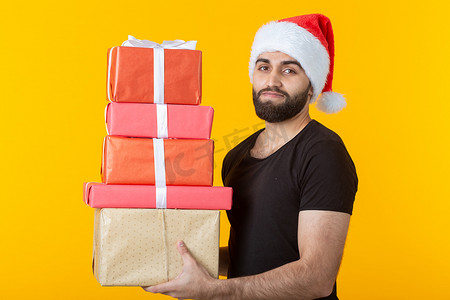心怀不满的年轻人戴着圣诞老人的帽子，留着胡子，手里拿着五个礼品盒，背景是黄色的。