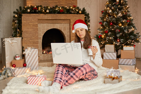 喝咖啡表情摄影照片_在家过圣诞节，在隔离期间庆祝寒假，面带悲伤表情的女士坐在地板上，膝上放着笔记本，手里拿着杯子喝咖啡或茶。