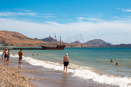 海洋上的轮船摄影照片_游客沿着黑海的卵石滩漫步，码头上停着一艘轮船。