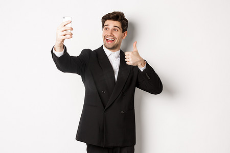 帅哥在新年派对上自拍、穿着西装、在智能手机上拍照并竖起大拇指、站在白色背景下的肖像