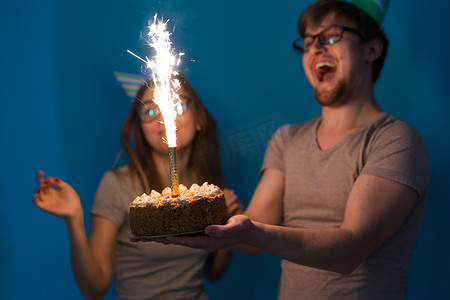 快乐的年轻夫妇迷人的男人和戴着纸帽的可爱女孩做傻脸，手里拿着一个站在蓝色背景上的生日蛋糕。
