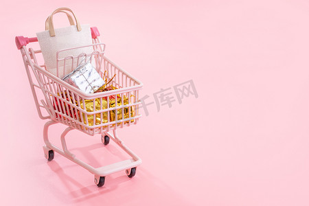 年度销售购物季概念 — 淡粉色背景中突显的装满纸袋礼品的迷你粉色购物车手推车、空白复印空间、特写