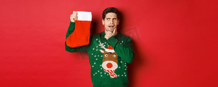 穿着绿色毛衣、体贴的帅哥的肖像，拿着圣诞袜思考，猜测里面是什么礼物，站在红色背景上