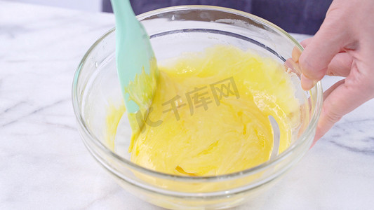 蛋黄摄影照片_用绿色橡胶刮刀搅拌工具将蛋黄混合到蛋糕面糊中，搅拌至光滑，并在玻璃碗中充分混合，特写，生活方式