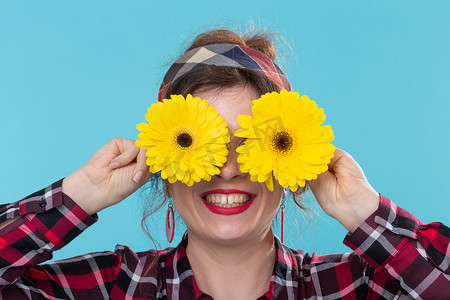 穿着红色格子衬衫、眼睛上挂着黄色花朵、在蓝色背景下摆出姿势的特写正面微笑的年轻女子。