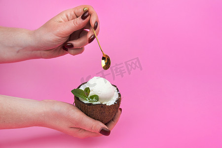 香草冰淇淋球放在新鲜椰子的一半里，手里拿着薄荷叶装饰，粉色背景上放着勺子