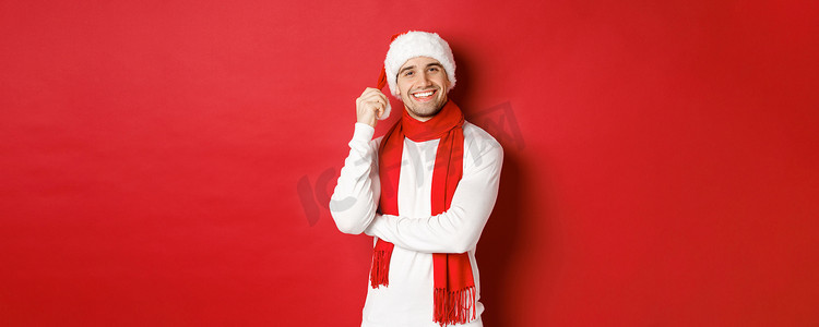 快乐男人享受圣诞假期的画像，戴着圣诞帽和围巾，快乐地微笑，站在红色背景上