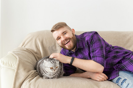 时髦的留着胡子的时髦青年抚摸着他坐在沙发上的美丽的灰色苏格兰折耳猫。
