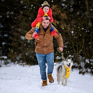 雪地里散步摄影照片_戴着黄色围巾的哈士奇与主人和他的女儿，在冬季的森林里散步。