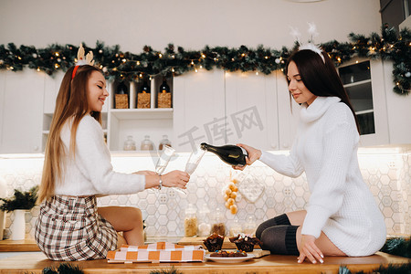 家庭环境摄影照片_两个女孩在舒适的家庭环境中，在厨房里为圣诞节倒香槟。