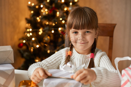 扎带摄影照片_女孩坐在客厅的桌子旁收拾礼物盒，带着迷人的微笑看着相机，扎着辫子，穿着白色毛衣，背景是圣诞树。