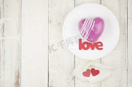 心形的花瓣摄影照片_桌上放着爱字和白色花瓣玫瑰，上面放着心形的红色蛋糕。