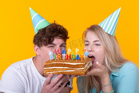 戴着纸帽的怪异年轻夫妇女孩和男孩的特写想咬掉一块祝贺蛋糕。