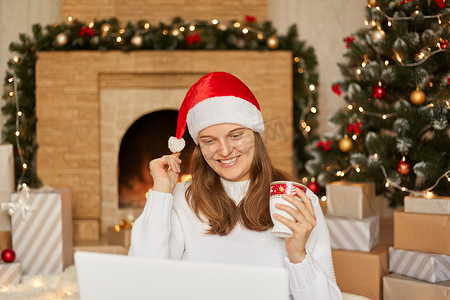 年轻女子用笔记本电脑坐在树前的地板上，用快乐的表情从圣诞老人的帽子上摸着她的绒球，祝贺她亲密的人过圣诞节。