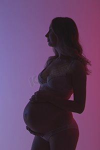 紫色灯光摄影照片_在美丽的霓虹灯紫色灯光下，穿着内衣的漂亮年轻孕妇在工作室里愉快地抚摸着她的肚子。