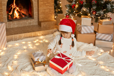 可爱的小女孩坐在地板上柔软的地毯上，在家里打开圣诞礼物，看着盒子，拉着丝带打开礼物，穿着白色套头衫和红色圣诞帽。