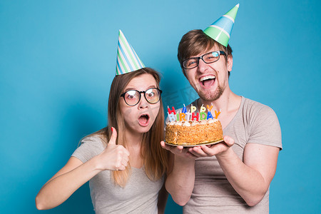 生日蛋糕海报摄影照片_快乐的年轻夫妇迷人的男人和戴着纸帽的可爱女孩做傻脸，手里拿着一个站在蓝色背景上的生日蛋糕。
