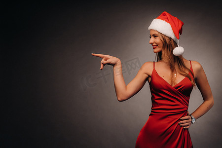 红色暗色摄影照片_一位身穿红色连衣裙、头戴圣诞帽的女士用手指指着黑色背景。新年微笑的女孩在深色背景中展示方向