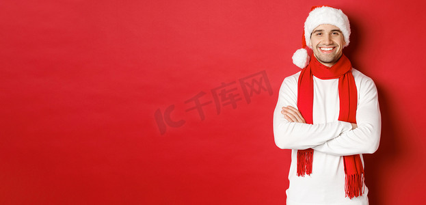 帅气的白种人，长着鬃毛，戴着圣诞帽、围巾和白色毛衣，双臂交叉在胸前，笑得开心，站在红色背景下