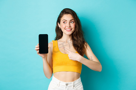 可爱的黑发女人用手指指着智能手机屏幕，向左看复制空间，站在蓝色背景下的夏季服装的肖像