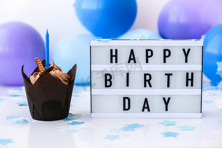 生日快乐摄影照片_带有文字“生日快乐”的灯箱和抽象的离焦模糊节日背景上的巧克力蛋糕蜡烛。