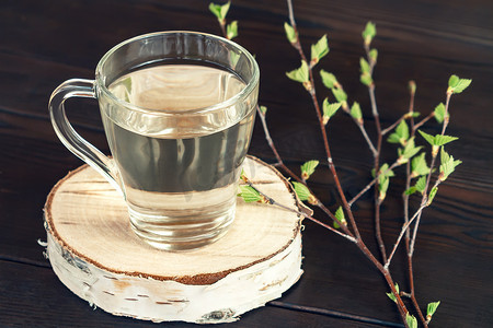 一张黑桌上的桦树汁放在一个玻璃杯里，旁边是一根有嫩叶的桦树枝