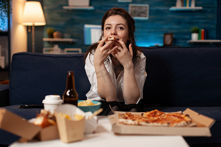 披萨盒摄影照片_看喜剧电影的女性肖像，吃着美味的外卖披萨片，在沙发上放松
