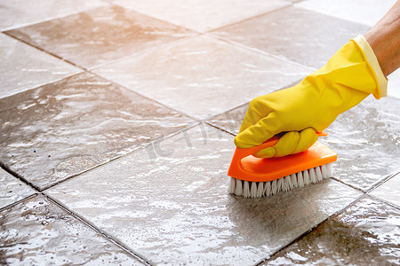 戴着黄色橡胶手套的手正用塑料地板刷用地板清洁剂擦洗瓷砖地板。