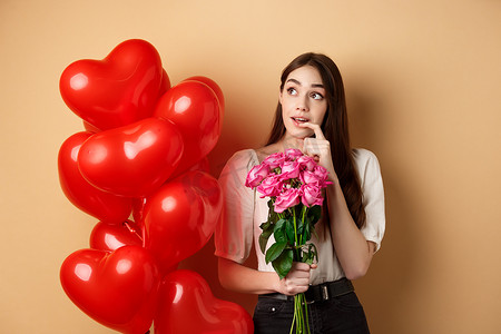 梦幻般的年轻女人手捧玫瑰花束，想着情人节的秘密仰慕者，看着左上角，咬着手指，站在红色浪漫气球旁