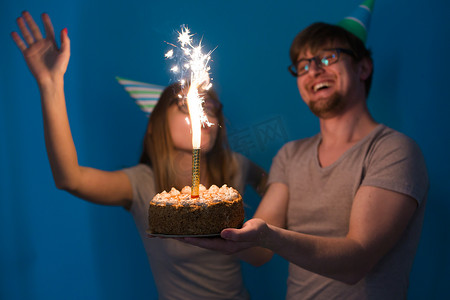 快乐的年轻夫妇迷人的男人和戴着纸帽子的可爱的女孩做出愚蠢的表情，手里拿着一个站在蓝色背景上的生日蛋糕。