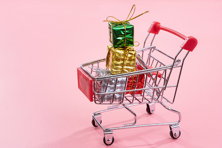 年度销售、圣诞购物季概念 — 迷你红色购物车手推车，里面装满了淡粉色背景中突显的礼盒，复制空间，特写