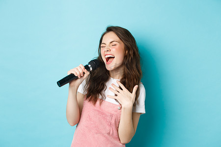美女歌手摄影照片_无忧无虑的美女表演歌曲，用麦克风热情地唱歌，唱卡拉 OK，站在蓝色背景上