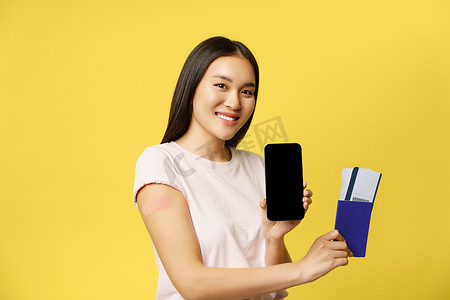 app展示界面摄影照片_微笑的亚洲女孩接种疫苗，展示护照票和智能手机屏幕、应用程序界面，站在黄色背景上