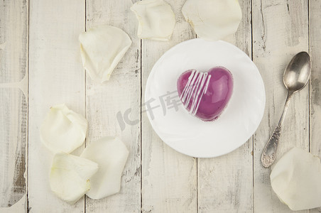 心形玫瑰花瓣摄影照片_桌上放着爱字和白色花瓣玫瑰，上面放着心形的红色蛋糕。