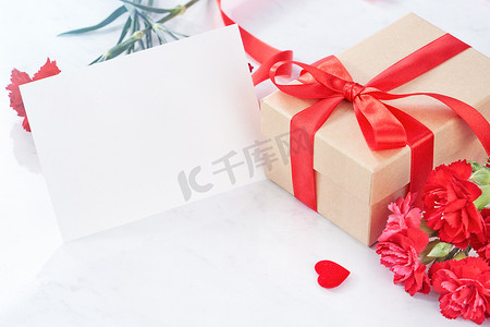 母亲节问候设计理念 — 美丽盛开的红色康乃馨，白色大理石桌背景上带丝带的礼盒，特写，复制空间。