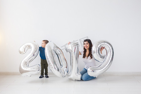 新年、庆祝和节日概念 — 母亲和儿子坐在 2019 年标志附近，用银气球在白色房间背景中迎接新年
