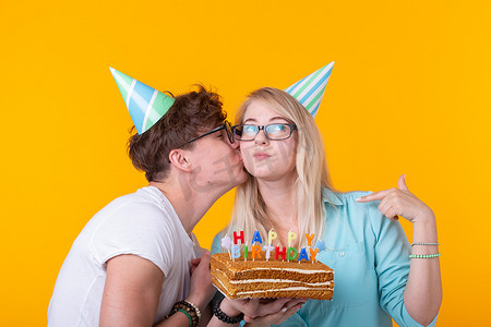 节日蓝色海报摄影照片_一对年轻迷人的男人和戴着纸帽的可爱女孩做傻脸，手里拿着蛋糕，黄色背景上刻着生日字样。