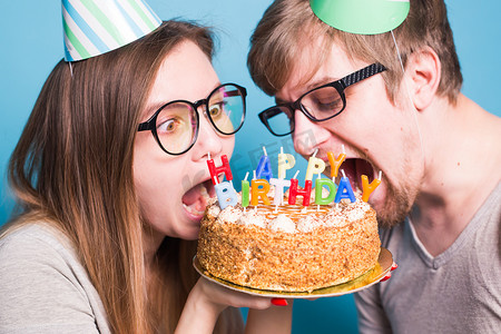 海报祝贺摄影照片_戴着纸帽的怪异年轻夫妇女孩和男孩的特写想咬掉一块祝贺蛋糕。