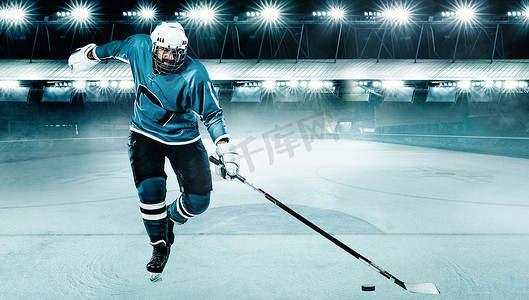 拿着棍子摄影照片_冰球运动员戴着头盔和手套在体育场上拿着棍子。