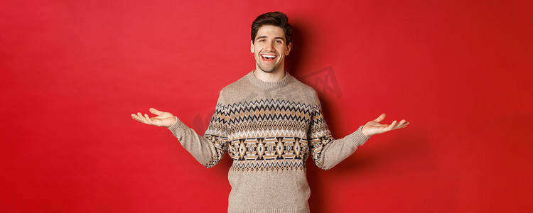 一双手拿东西摄影照片_庆祝新年假期的快乐帅哥的画像，穿着圣诞毛衣，双手横向展开，微笑着，拿着一些东西在复印空间，站在红色背景上