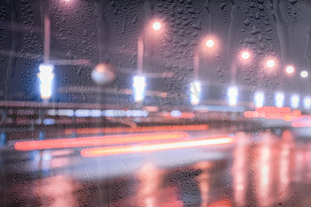 下雨的夜摄影照片_在被雨水覆盖的窗户后面的夜城里，移动的汽车和灯笼的模糊灯光反射在潮湿的沥青上。