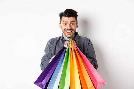 站在白色背景下，兴奋的微笑男子拿着彩色购物袋，对商店里的折扣感到高兴