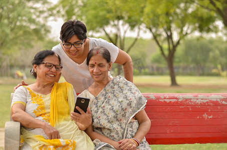 印度新德里，一位年轻的印度女孩和婆婆坐在公园的红色长椅上，是年长的优雅女性，她们坐在手机屏幕上闲聊和微笑。