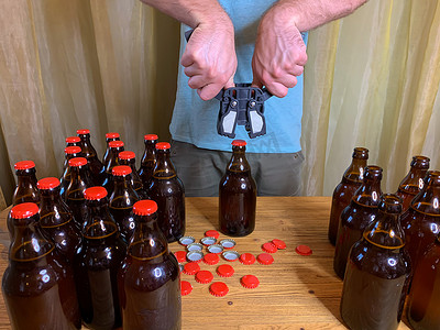 啤酒瓶啤酒盖摄影照片_在家里酿造精酿啤酒，男人在木桌上用塑料封盖器关闭棕色玻璃啤酒瓶，上面有红色皇冠盖。