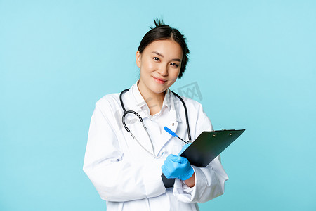女医生的画像，身着医疗制服的亚洲医生，写下病人信息，站在蓝色背景下