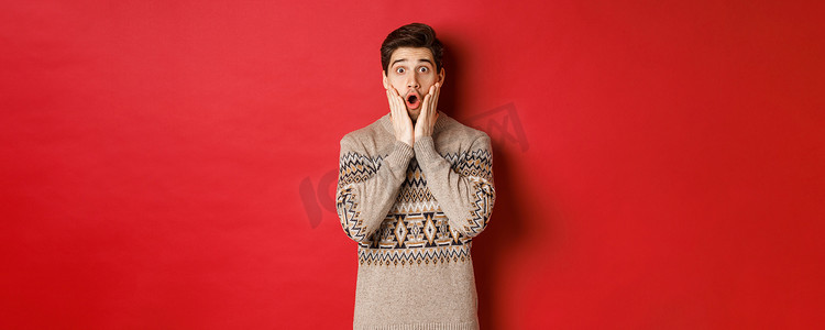 炫酷背景红色摄影照片_惊讶的帅哥对酷炫的新年促销活动做出反应，惊讶地喘着粗气，穿着圣诞毛衣，站在红色背景上