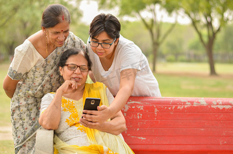 印度新德里，一位年轻的印度女孩和婆婆坐在公园的红色长椅上，是年长的优雅女性，她们坐在手机屏幕上闲聊和微笑。